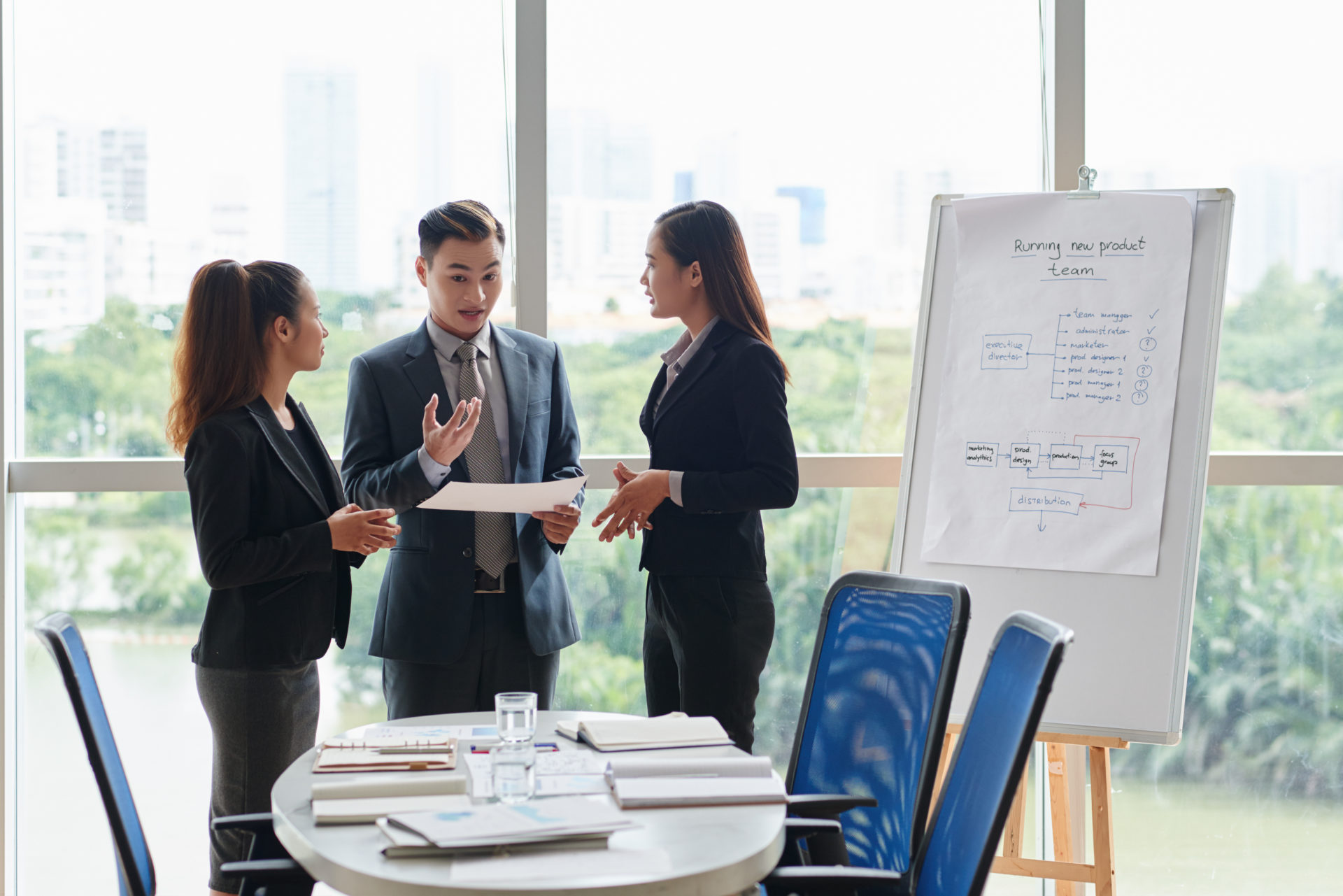 Handsome Asian businessman with stylish haircut giving a piece of advice to his pretty female colleagues while standing at panoramic window of spacious meeting room